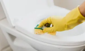 Ręka w żółtej rękawiczce czyści deskę sedesową gąbką. Dowiedz się, jak usunąć zapach chloru z pomieszczenia, by cieszyć się świeżością.