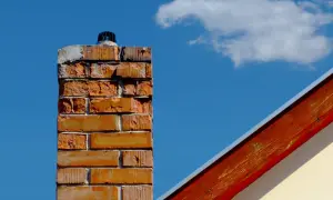 Woda w kominie po deszczu. Ceglany komin z metalową nakładką na tle błękitnego nieba i fragmentu dachu.