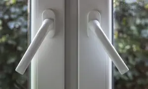 Dwie białe klamki okienne, gotowe do wymiany, na tle zieleni za szybą.