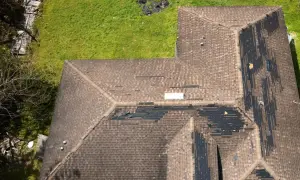Widok z góry na dach z uszkodzeniami pokrycia, widoczne czarne plamy i zerwane gonty. Prawdopodobne przeciekanie dachu przy kominie.
