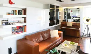 Nowoczesna zabudowa wyczystki z białymi frontami, półkami z książkami i brązową kanapą. Obok fortepian i designerska lampa.