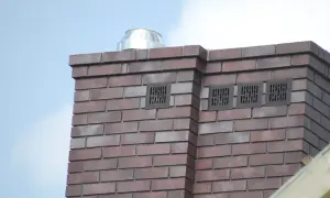 Nowy komin z cegły klinkierowej, z metalowym daszkiem i kratkami wentylacyjnymi, zgodny z normą kominową.