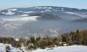 Mgła unosi się nad zimową doliną, ukazując sporą ilość pyłów zawieszonych w powietrzu. W oddali widać ośnieżone wzgórza i lasy.