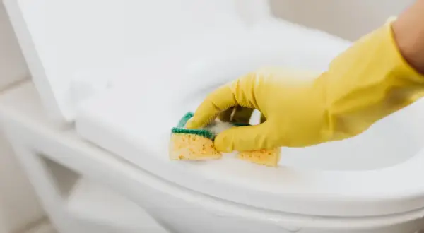 Ręka w żółtej rękawiczce czyści deskę sedesową gąbką. Dowiedz się, jak usunąć zapach chloru z pomieszczenia, by cieszyć się świeżością.
