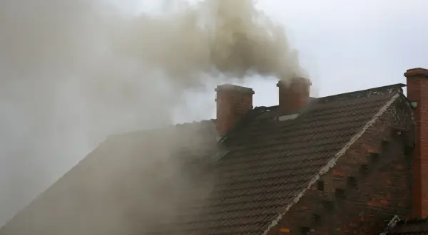 Gęsty, żółty dym z komina unosi się nad brązowym dachem.