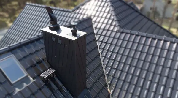 Czarny, nowoczesny komin na dachu z dachówki ceramicznej. Obok okno dachowe i stopień kominiarski.