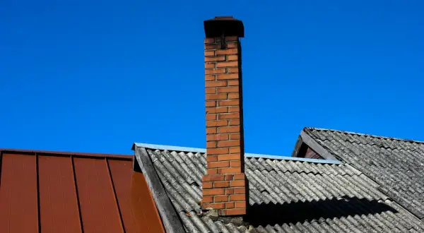 Ceglany komin na dachu, z którego wydobywa się sadza, na tle błękitnego nieba.