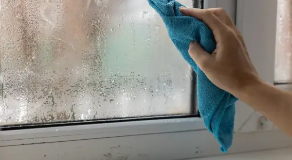 Ręka z niebieską ściereczką wyciera zaparowane okno. Ściereczka skutecznie pochłania wilgoć, przywracając przejrzystość szyby.