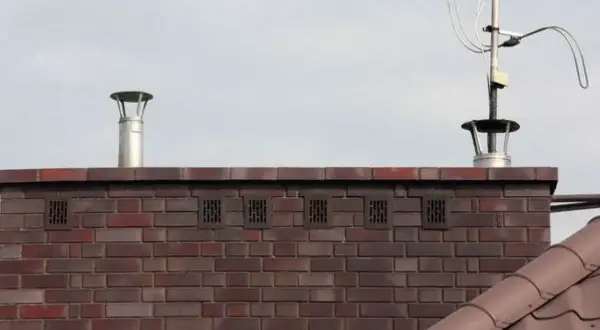 Zakończenie komina wentylacyjnego z metalową nasadką i kratkami wentylacyjnymi w ceglanej ścianie. Obok widoczna antena telewizyjna.
