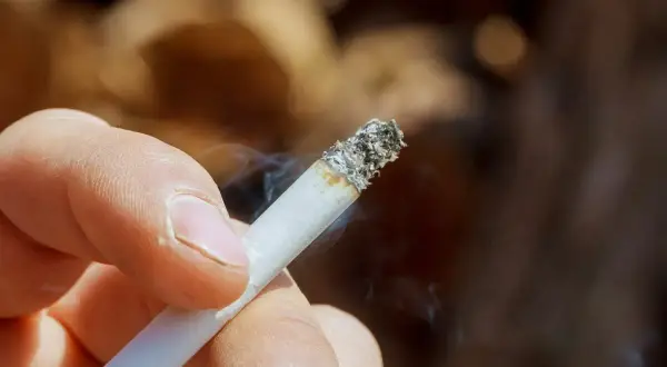 Dłoń trzymająca zapalonego papierosa, z którego unosi się dym. Zastanawiasz się, jak usunąć zapach papierosów ze ścian?