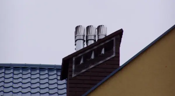 Trzy nasady kominowe na ceglanym kominie, świadczące o prawidłowym montażu nasady kominowej. Obok dach z niebieską dachówką.
