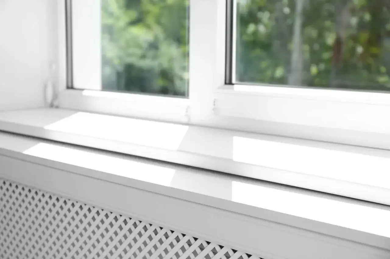Biały parapet pod okno, z widocznym fragmentem grzejnika, odbija światło słoneczne.