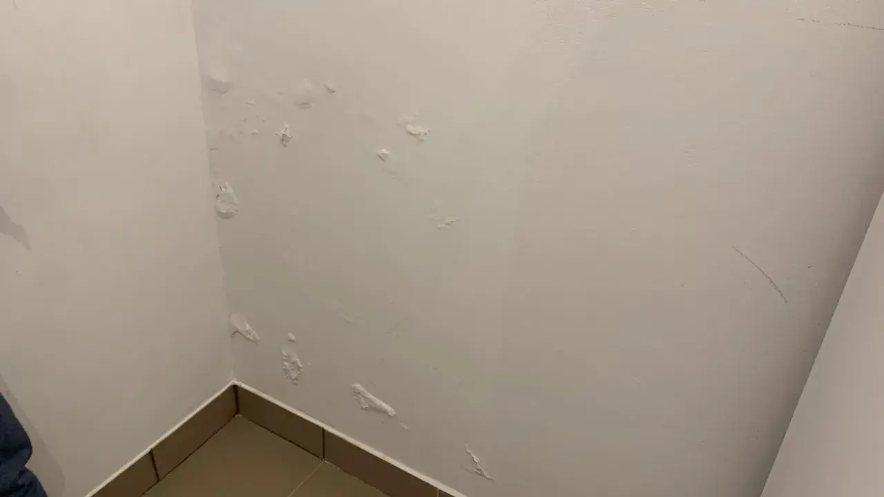 Widoczne pęcherze i łuszcząca się farba na białej, mokrej ścianie w rogu pokoju, wskazujące na problem z wilgocią.