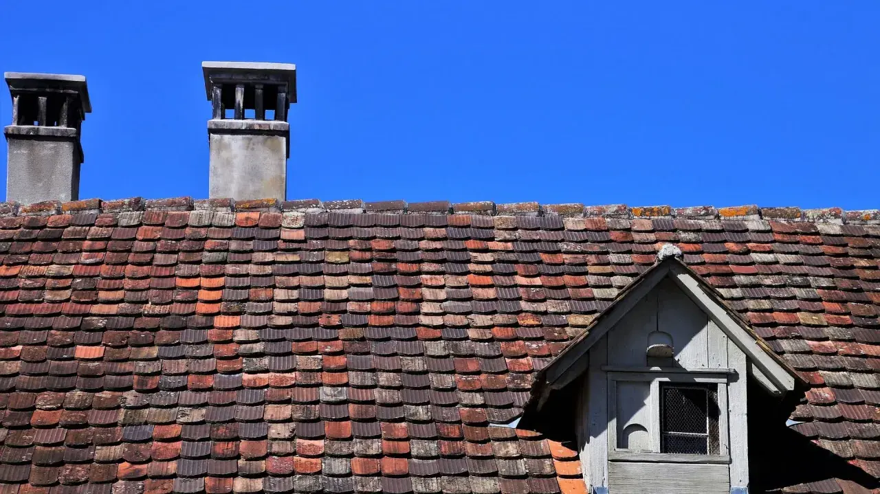 Dwa kominy na dachu pokrytym starymi dachówkami, wymagające naprawy komina z cegły.