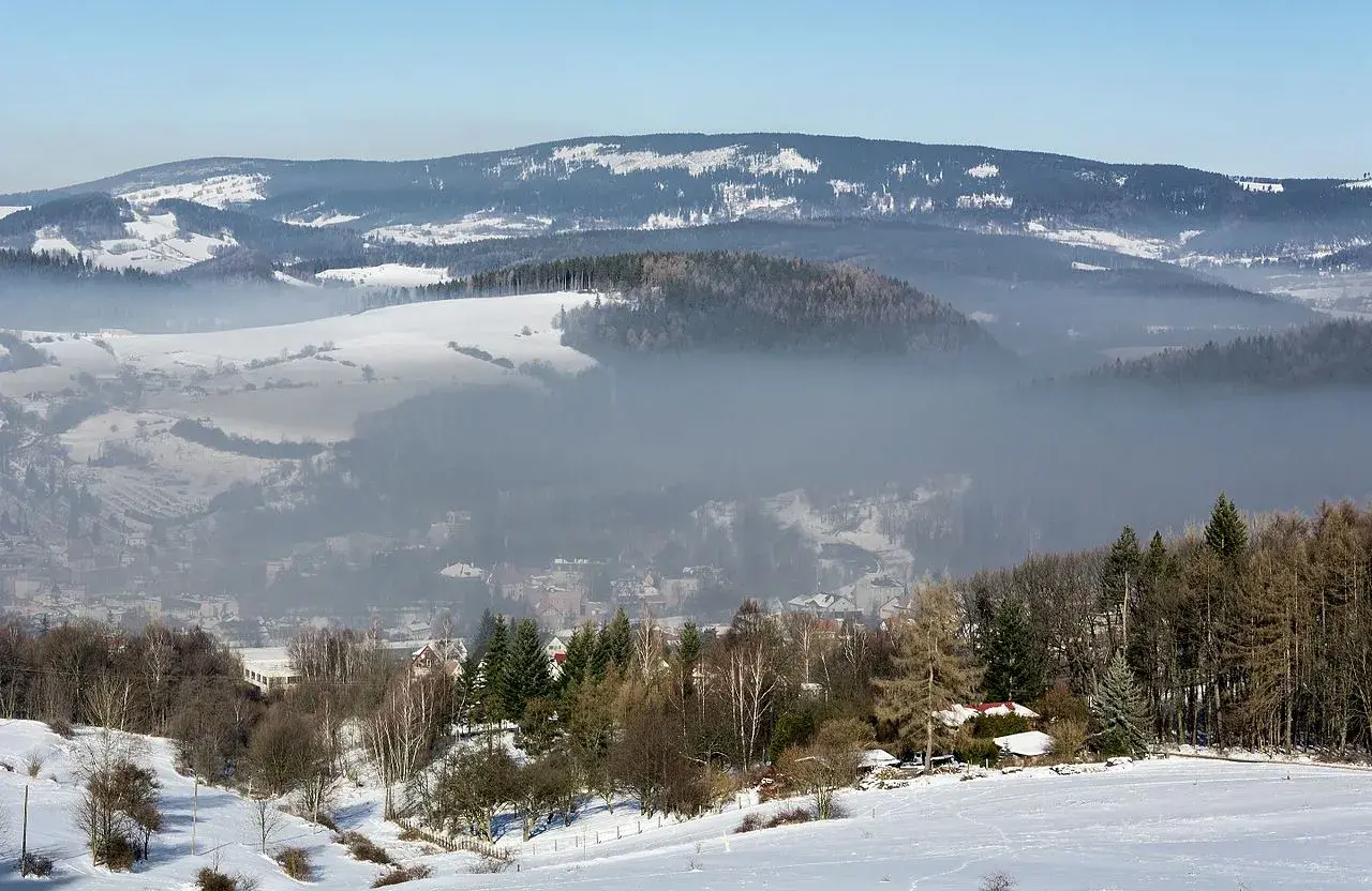 Mgła unosi się nad zimową doliną, ukazując sporą ilość pyłów zawieszonych w powietrzu. W oddali widać ośnieżone wzgórza i lasy.