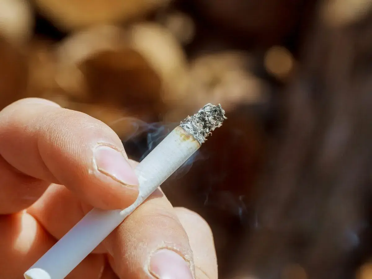 Dłoń trzymająca zapalonego papierosa, z którego unosi się dym. Zastanawiasz się, jak usunąć zapach papierosów ze ścian?