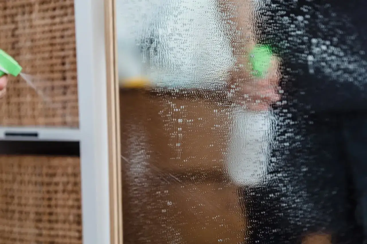 Ręka z zielonym spryskiwaczem aplikuje płyn na zaparowane okno. Dowiedz się, czym zmyć grunt z okna, by uzyskać idealną czystość.