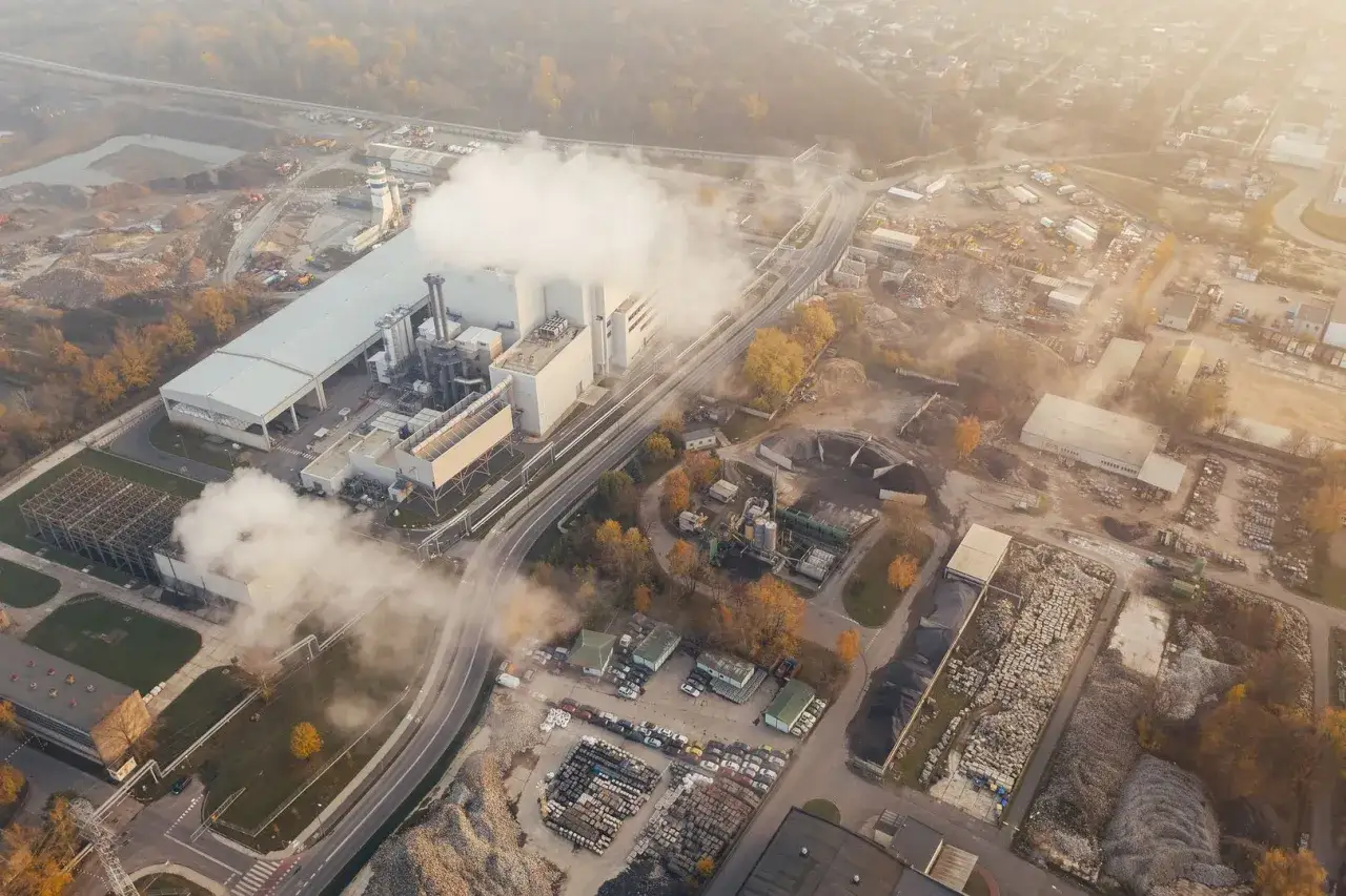 Widok z lotu ptaka na zakład przemysłowy z unoszącą się parą, która wpływa na jakość powietrza. Widać drogi, składy materiałów i budynki.