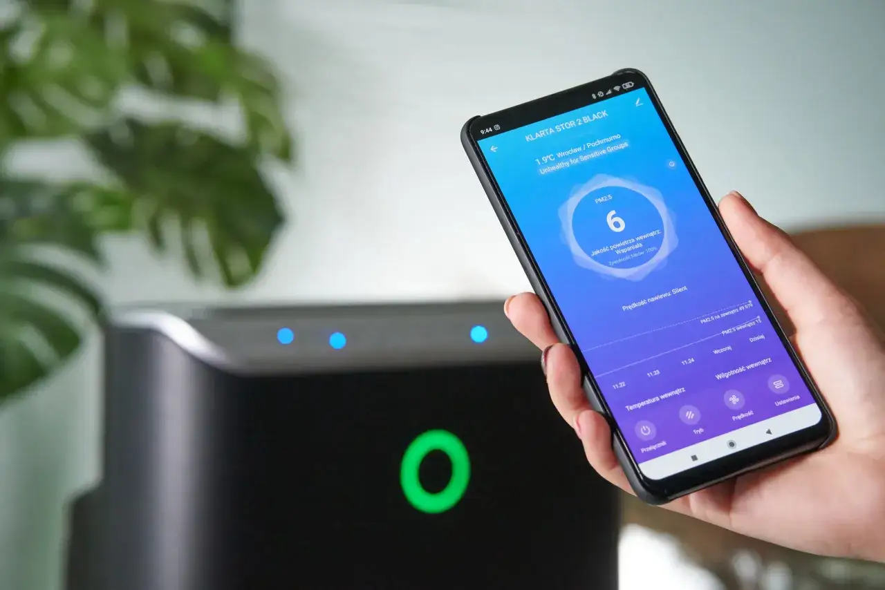 Dłoń trzyma smartfon z aplikacją monitorującą jakość powietrza. Wskaźnik PM2.5 pokazuje 6, co oznacza wspaniałą jakość. Urządzenie działa zgodnie z lzo normy.