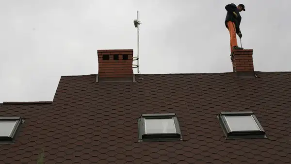 Mężczyzna w pomarańczowych spodniach stoi na kominie i czyści go szczotką.