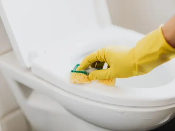 Ręka w żółtej rękawiczce czyści deskę sedesową gąbką. Dowiedz się, jak usunąć zapach chloru z pomieszczenia, by cieszyć się świeżością.