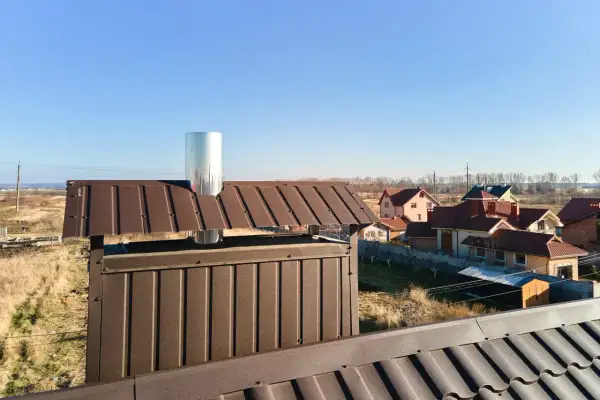 Nowy montaż rur kominowych na dachu z brązowej blachy. W tle domy i błękitne niebo.