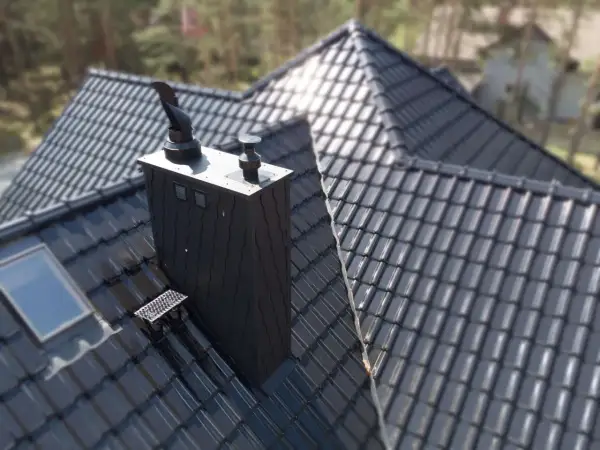 Czarny, nowoczesny komin na dachu z dachówki ceramicznej. Obok okno dachowe i stopień kominiarski.