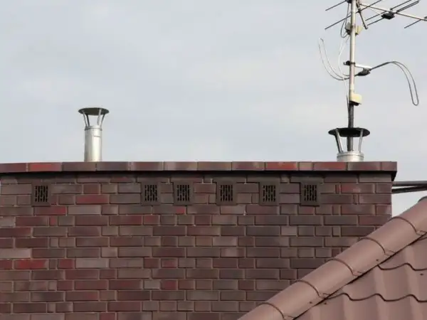 Zakończenie komina wentylacyjnego z metalową nasadką i kratkami wentylacyjnymi w ceglanej ścianie. Obok widoczna antena telewizyjna.