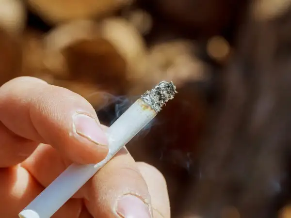 Dłoń trzymająca zapalonego papierosa, z którego unosi się dym. Zastanawiasz się, jak usunąć zapach papierosów ze ścian?