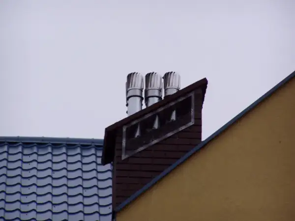 Trzy nasady kominowe na ceglanym kominie, świadczące o prawidłowym montażu nasady kominowej. Obok dach z niebieską dachówką.